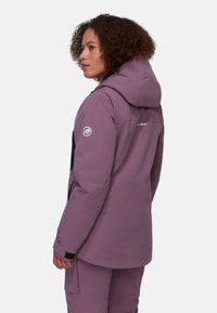 Lila wasserdichte Jacke mit Kapuze, ausgestattet mit schwarzen Akzenten an den Schultern und unteren Ärmeln sowie einem Logo am Oberarm. Strukturierter Stoff.