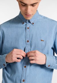 Lichtblauw spijkerhemd met button-down kraag en bruine knopen, voorzien van een klein geborduurd hondenontwerp op de linkerborst.
