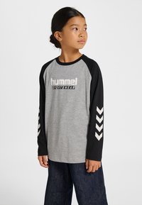 Grå långärmad tröja med svarta raglansärmar, med en "hummel"-logotyp och vitt chevronmönster på ärmarna. Matchad med mörka jeans.