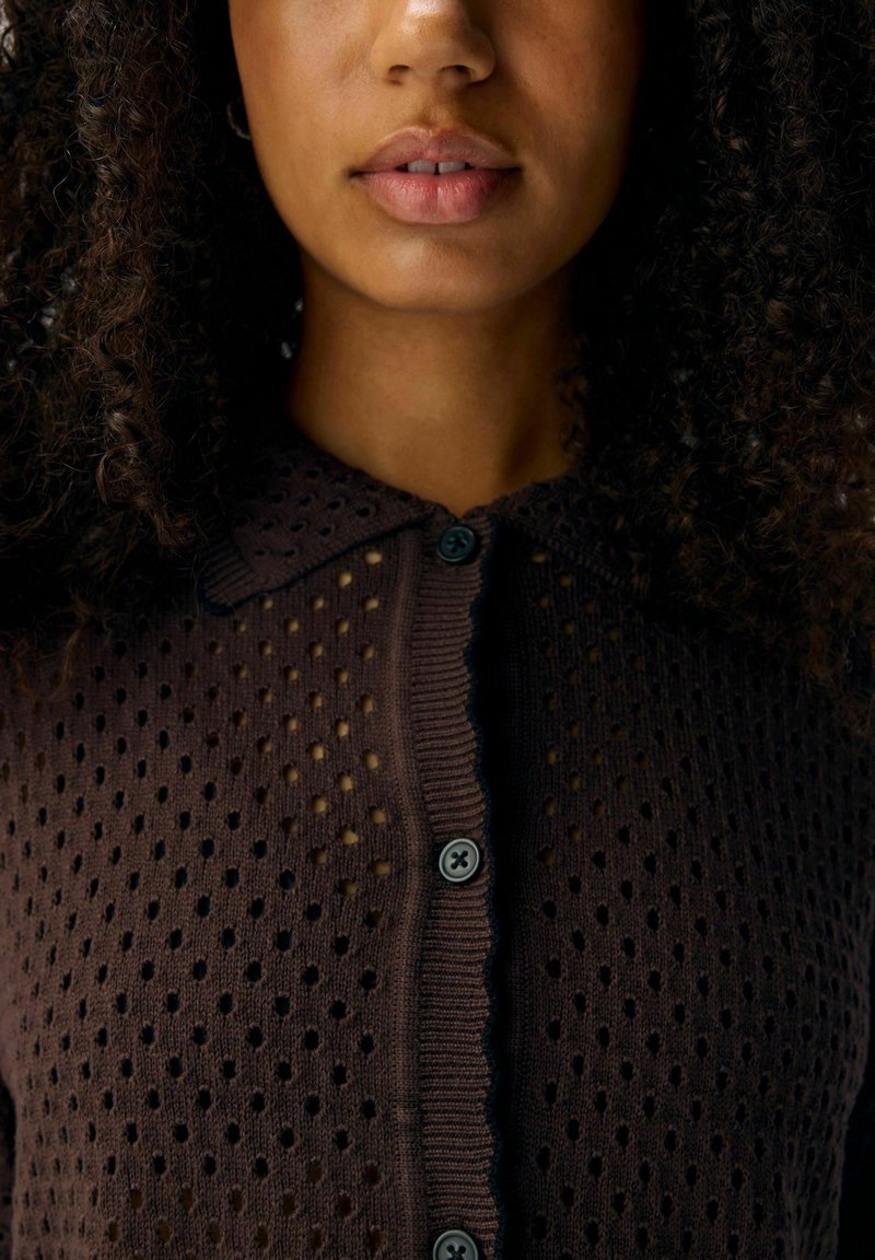 Kobieta z kręconymi włosami, ubrana w ciemno-brązowy sweter z guzikami o teksturowanym, kropkowanym wzorze i kołnierzyku.