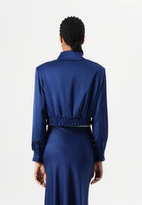 Blusa corta blu navy con maniche lunghe, cintura elasticizzata, colletto raffinato e texture liscia e lucida. È visibile una gonna lunga complementare.