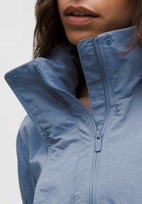 Vaaleansininen takki, jossa on korkea kaulus ja edessä vetoketju sekä nepparit. Kangas näyttää sileältä, mutta siinä on hieman ryppyinen rakenne.