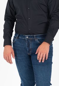 Camisa negra de botones combinada con jeans de mezclilla azul oscuro que cuentan con un bolsillo frontal y cierre de botón metálico. La tela tiene una textura suave.