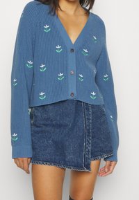 Blå stickad cardigan med V-ringning, prydd med gröna och vita blommönster, knappfront och ribbad textur. Matchad med en denimkjol.