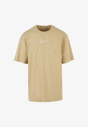 Muškarac s kratkom plavom kosom nosi bež oversize majicu, crne cargo hlače i bež tenisice, stoji s jednom rukom na glavi.