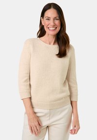 Beiger Strickpullover mit rundem Ausschnitt, Dreiviertelärmeln sowie gerippten Bündchen und Saum, getragen mit hellen Hosen.
