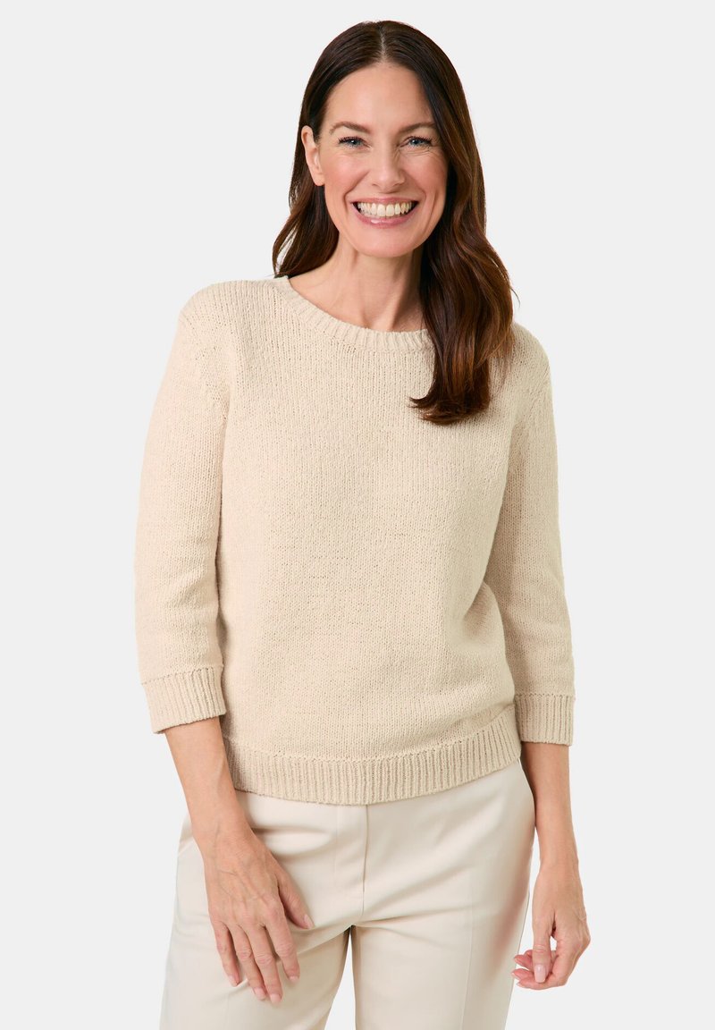 Beiger Strickpullover mit rundem Ausschnitt, Dreiviertelärmeln sowie gerippten Bündchen und Saum, getragen mit hellen Hosen.
