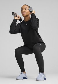 Black athletic sweatshirt and leggings with Puma logo; individual holding dumbbells, wearing blue and white sneakers, demonstrating a squat.