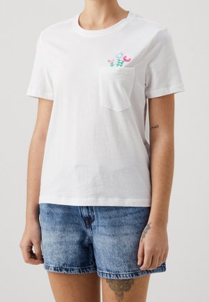 Osoba w białej koszulce z krótkim rękawem z kolorowymi haftowanymi kwiatami na kieszeni, w połączeniu z niebieskimi szortami dżinsowymi oraz widocznymi tatuażami na ramionach i nodze.