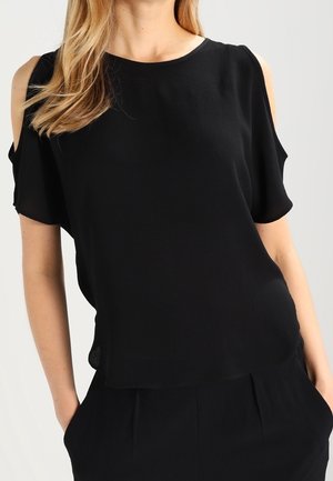Femme portant un chemisier noir épaules dénudées avec manches courtes et un pantalon noir, les mains dans les poches.