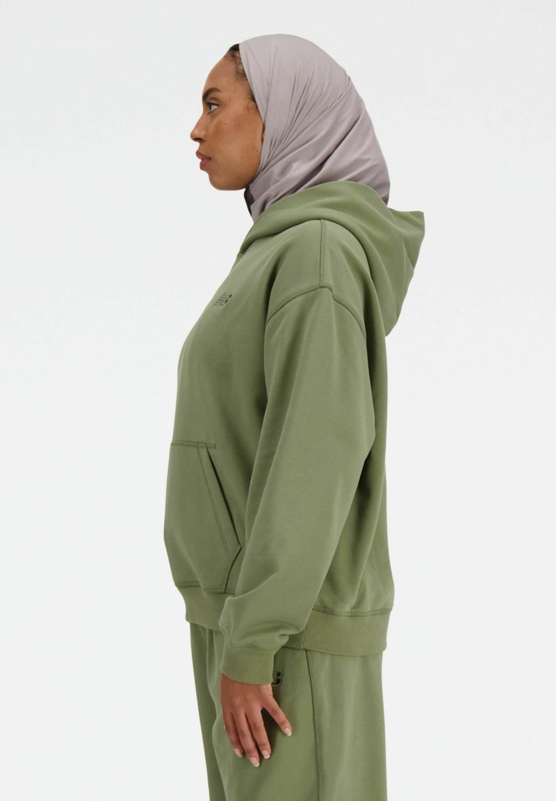 Vrouw in een lichtgrijze hijab en een olijfgroen hoodie met bijpassende broek, staande in profiel tegen een effen witte achtergrond.