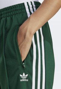Vihreät urheiluhousut vetoketjullisella taskulla. Sivussa kolme valkoista raitaa ja edessä Adidas-logo. Pehmeä kangaspinta.