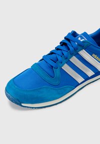 Sapatilha atlética azul com materiais em camurça e malha, apresentando três riscas brancas, uma sola texturizada e uma língua azul com detalhes de logotipo.