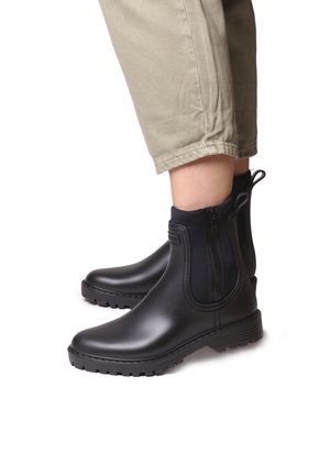 Black rubber ankle boots with a smooth finish, zip closure, and textured sole, paired with light khaki pants.
