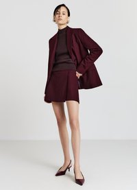 Burgunderroter Blazer mit feinen Nadelstreifen, passender kurzer Rock und eng anliegender Rollkragenpullover. Abgerundet wird das Outfit durch spitze Slingback-Pumps. Glatte Textur.