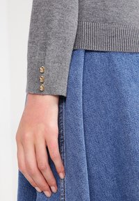 Pull gris en maille avec ourlet côtelé et trois boutons dorés sur le poignet, associé à une jupe en denim. Accent sur le contraste de texture et de couleur.