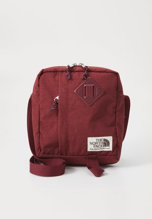 Sac bandoulière carré marron avec fermeture éclair frontale, sangle réglable, patch en forme de losange et patch logo The North Face en bas à droite.