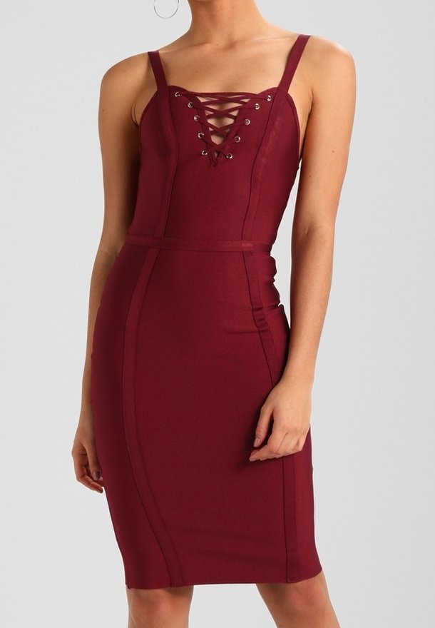 Femme portant une robe bordeaux ajustée avec de fines bretelles et un détail à lacets sur le devant, sur un fond gris uni.