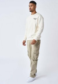 Sweatshirt beige avec texte brodé, associé à un pantalon cargo kaki avec poches latérales et des baskets blanches. Texture de tissu lisse.
