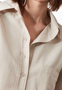 Primo piano di una persona che indossa una camicia beige abbottonata con taschino, mostrando parte del collo e un orecchino.