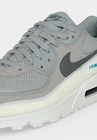 Γκρι αθλητικό παπούτσι Nike Air Max με υφές από δίχτυ και σουέτ, γκρι κορδόνια, μαύρο λογότυπο Nike και λευκή σόλα με ορατή αερόσολα.
