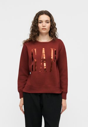 Tânără cu păr creț poartă un hanorac bordo cu litere mari reflectorizante "GAP" pe față și pantaloni largi negri.