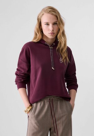 Joven con cabello rubio largo y ondulado, vestida con sudadera con capucha granate y pantalones grises con cordón, con las manos en los bolsillos sobre un fondo liso.