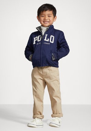 Giubbotto bomber blu navy con logo "POLO", abbinato a pantaloni beige. Il bambino indossa sneakers bianche, con una camicia a quadri visibile sotto.