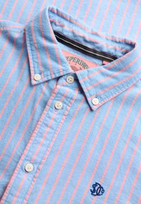 Camicia azzurra con bottoni, righe verticali rosa, colletto con bottoni, bottoni bianchi e un piccolo logo ricamato blu scuro vicino all'orlo.