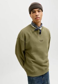 Oliwkowy sweter z ściągaczami przy mankietach i dolnym brzegu, z małym czarnym logo. Nosi się go na niebieskiej chustce w wzór paisley i w zestawie z niebieskimi dżinsami.