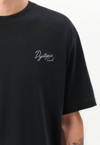 Svart T-skjorte med hvit skrifttype "Dystopia Sounds." på brystet, båret av en person med en synlig tatovering på underarmen.