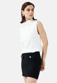 Top de cuello alto sin mangas blanco combinado con una minifalda negra texturizada que presenta dos bolsillos frontales y acentos de botones plateados.