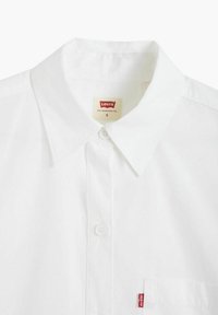 Chemise blanche en coton avec col et fermeture à boutons, arborant une petite étiquette rouge Levi's sur la poche gauche. Texture lisse et design classique.