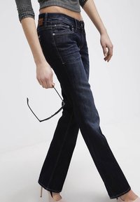 Jeans blu scuro con vestibilità a gamba dritta, caratterizzati da accessori in argento, una toppa in pelle marrone sulla cintura e dettagli di cucitura visibili.