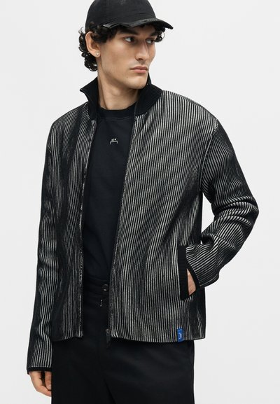 Άνδρας που φοράει μαύρο καπέλο, μαύρο και γκρι κατακόρυφη ριγέ ζακέτα με φερμουάρ, μαύρο πουκάμισο και σκοτεινό παντελόνι, κοιτάζει προς τα πλάγια με το ένα χέρι στην τσέπη.