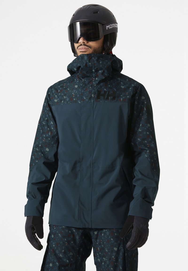 Waterproof ski jacket with a dark teal body, patterned sleeves, high collar, and logo. Matched with black gloves, showcasing a functional design.