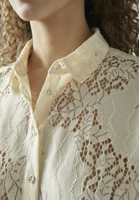 Blonde bluse i lys beige med blomstermønster, der har krave med knaplukning og tekstureret stof, der fremhæver den intrikate blonde detaljering.