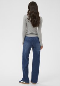 Femme debout de dos, portant un haut à manches longues gris, un jean bleu droit et des baskets bleues avec des rayures blanches.