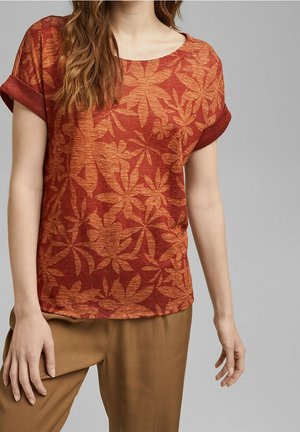 Mujer con una camiseta de manga corta color óxido con un patrón floral naranja y pantalones marrones sobre un fondo gris liso.