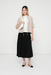 Kurzärmelige, hellbeige, cropped Strickjacke über einem weißen Oberteil, kombiniert mit einem schwarzen, plissierten Rock und flachen Schuhen mit schwarzen Akzenten.