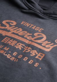 Sudadera con capucha de color gris oscuro que presenta un gran logo impreso en cobre con textura, que incluye el texto "Vintage SuperDry" y caracteres japoneses adicionales.