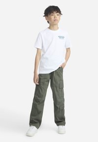 Λευκό βαμβακερό μπλουζάκι με πράσινο κείμενο και λογότυπο, συνδυασμένο με χακί cargo παντελόνι με τσέπες και μανσέτες. Λευκά αθλητικά παπούτσια ολοκληρώνουν το ντύσιμο.