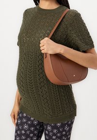 Frau in einem olivgrünen, gestrickten, kurzärmligen Pullover und schwarzen Blumenhosen, die eine braune lederne Schultertasche mit gebogener Form trägt.