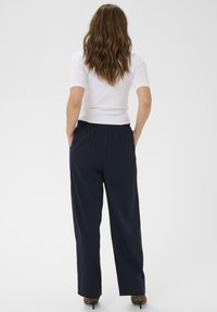 Ženska z valovitimi rjavimi lasmi stoji s hands in pockets, oblečena v belo kratkotrajno majico, modre ohlapne hlače in čevlje s tiskanjem leoparda, obrnjena stran.