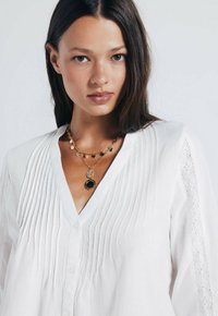 Hvid, letbluse med lodrette læg, dyb V-hals og blonde detaljer på ærmerne. Sammen med et lagdelt guldkæde med sorte accenter.