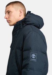 Marineblå pufferjakke med h�øj krave og hætte, der har et Timberland-logo på ærmet og en glat, blank overflade.