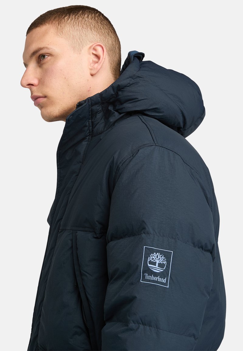 Marineblaue Steppjacke mit hohem Kragen und Kapuze, ausgestattet mit einem Timberland-Logopatch am Ärmel und einer glatten, glänzenden Oberfläche.
