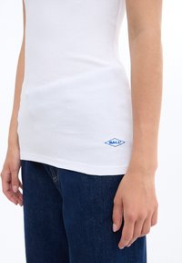Camiseta sin mangas blanca con textura acanalada que presenta un logotipo bordado azul en el dobladillo, combinada con jeans de mezclilla azul oscuro. Corte ajustado con una textura suave.