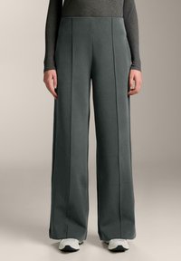 Breda byxor i mjuk grått tyg, med framfållar, hög midja och slät struktur. Matchas med vita sportskor.