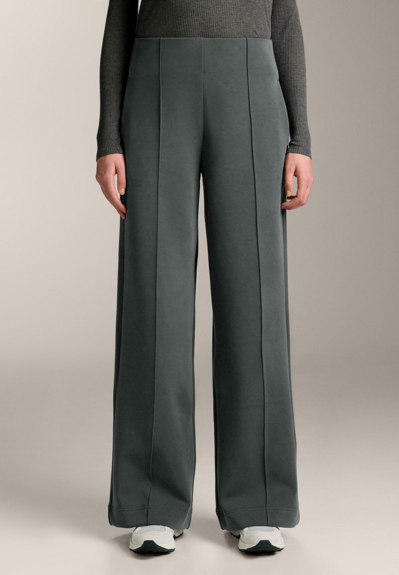 Breda byxor i mjuk grått tyg, med framfållar, hög midja och slät struktur. Matchas med vita sportskor.